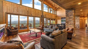 A spacious living area with wooden walls, large windows, and a stone fireplace. - 6BR for 26 | Pool, Hot Tub, Game Loft, & Mini Golf (Sevierville)