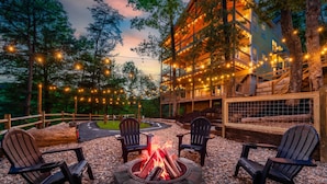 A cozy outdoor seating area with Adirondack chairs around a stone fire pit, surrounded by a wooden deck and string lights. - Summit Luxe Resort (Gatlinburg)