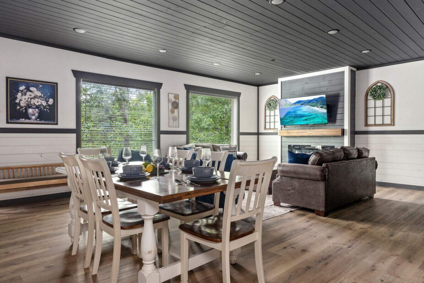 A dining area with a wooden table set for four, surrounded by white chairs. There is a grey sofa and a television mounted on the wall.
