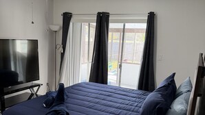 Ett sovrum med en säng, en tv, en takfläkt, ett fönster med gardiner och ett litet bord med en lampa och fjärrkontroller.