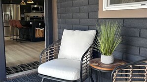 A modern living area with a black brick wall, a white cushioned chair, a round side table with a plant, and a view of a kitchen and dining area through an open door. - Downtown Ebony- 1.5 Miles from Whiskey Row (Prescott)