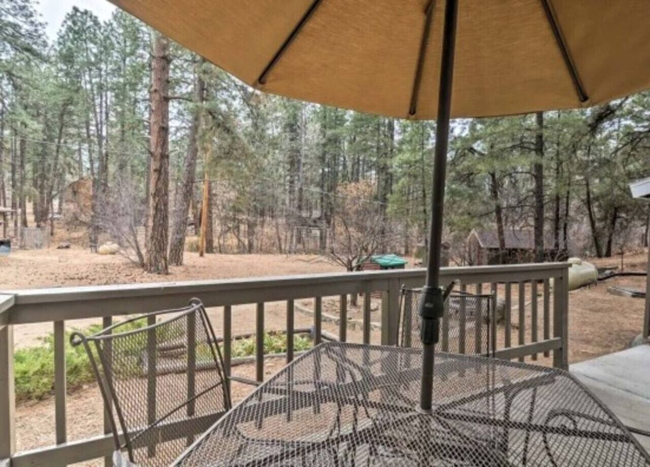 A covered outdoor area with a metal table and chairs, surrounded by a wooden deck and a forested background.