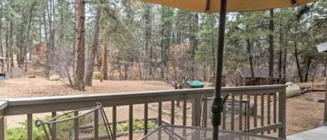 A covered outdoor area with a metal table and chairs, surrounded by a wooden deck and a forested background.