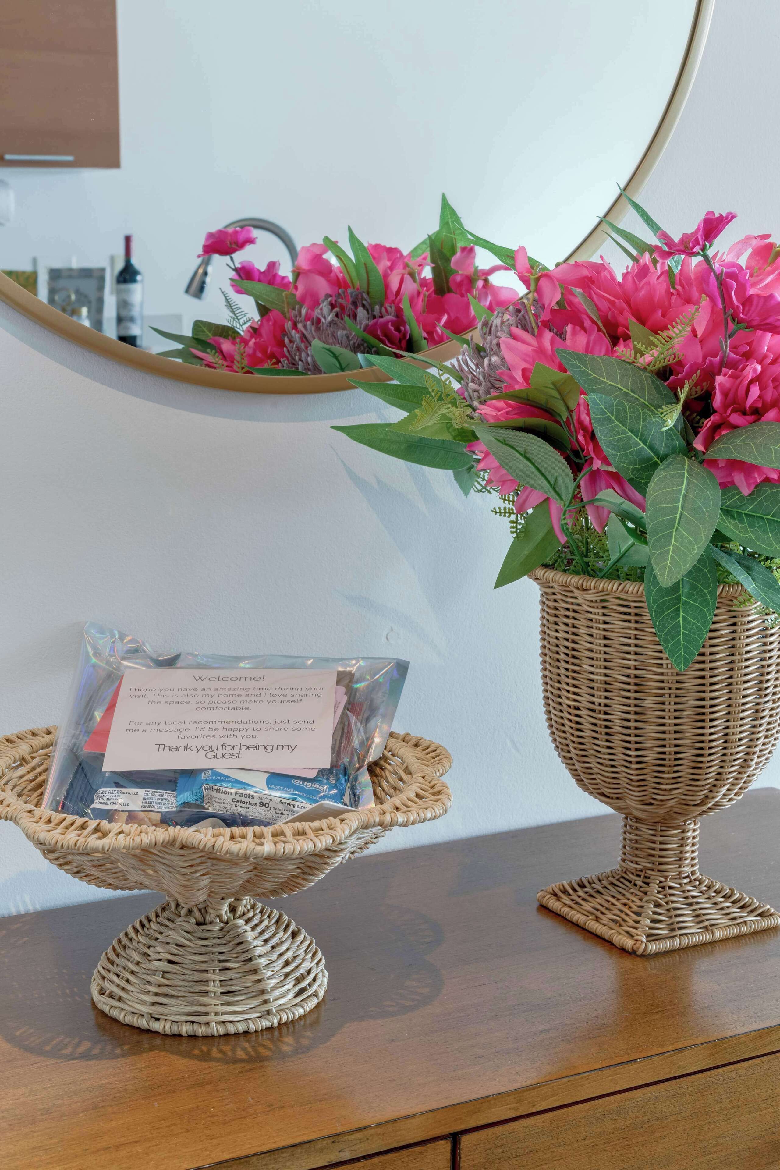 En fletkurv med et velkomstkort og en flevase med lyserøde blomster på et træbord.