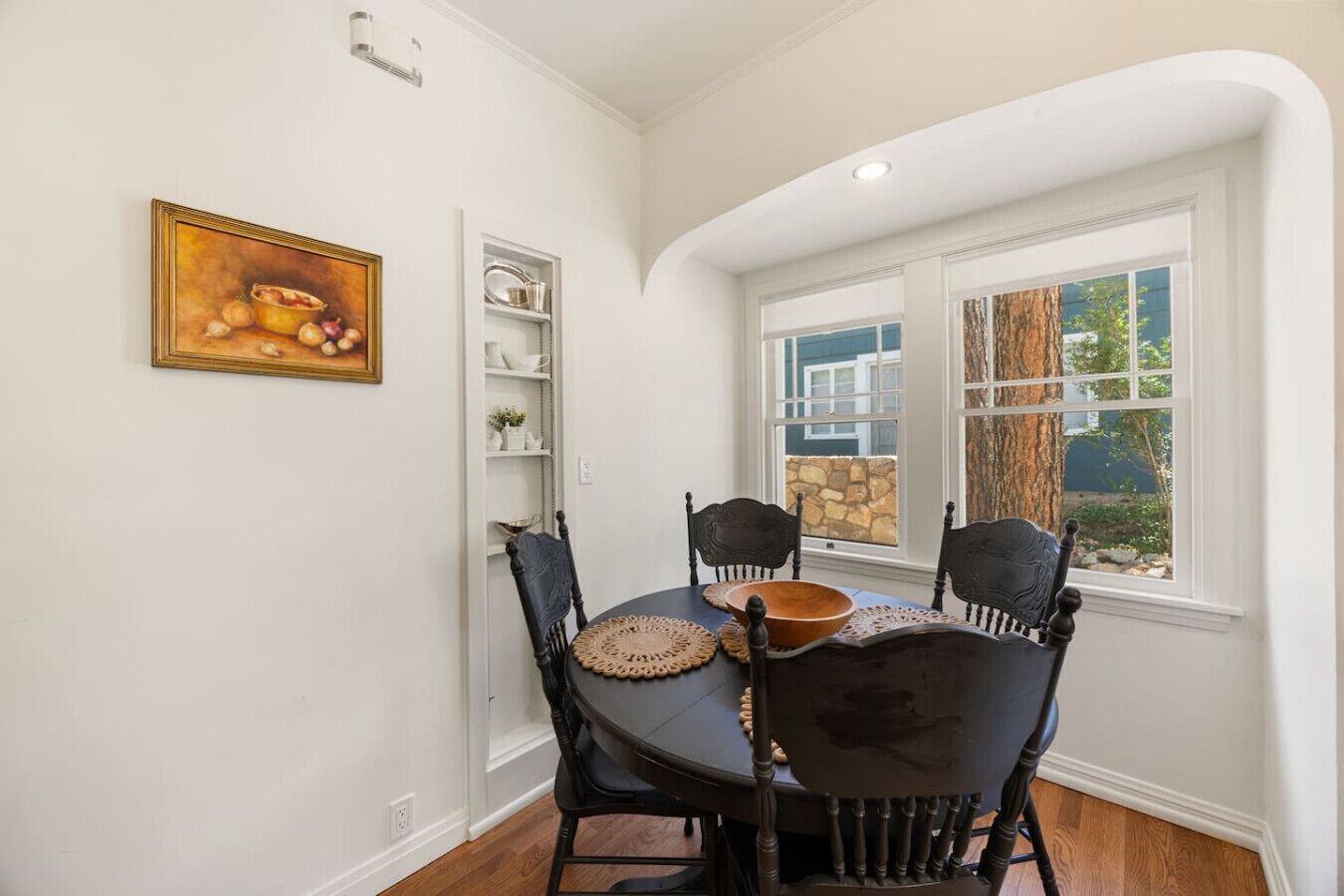 A dining area with a round table, four chairs, a bowl, and a shelf with dishes.