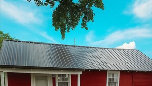 A red single-story house with a metal roof, a front porch, and a small garden area. - Peaceful 1-bed studio cottage in Gering perfect for relaxing stay (Gering)