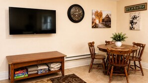 A living room with a TV, clock, paintings, a wooden table, chairs, and a shelf with board games. - Peaceful 1-bed studio cottage in Gering perfect for relaxing stay (Gering)