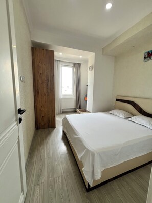 Habitación de hotel con una cama grande, piso de madera, una ventana con cortinas y una mesita de noche pequeña.