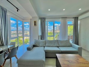 Una sala moderna con un sofá modular, una mesa de comedor y sillas. Amplias ventanas ofrecen vistas a la ciudad.