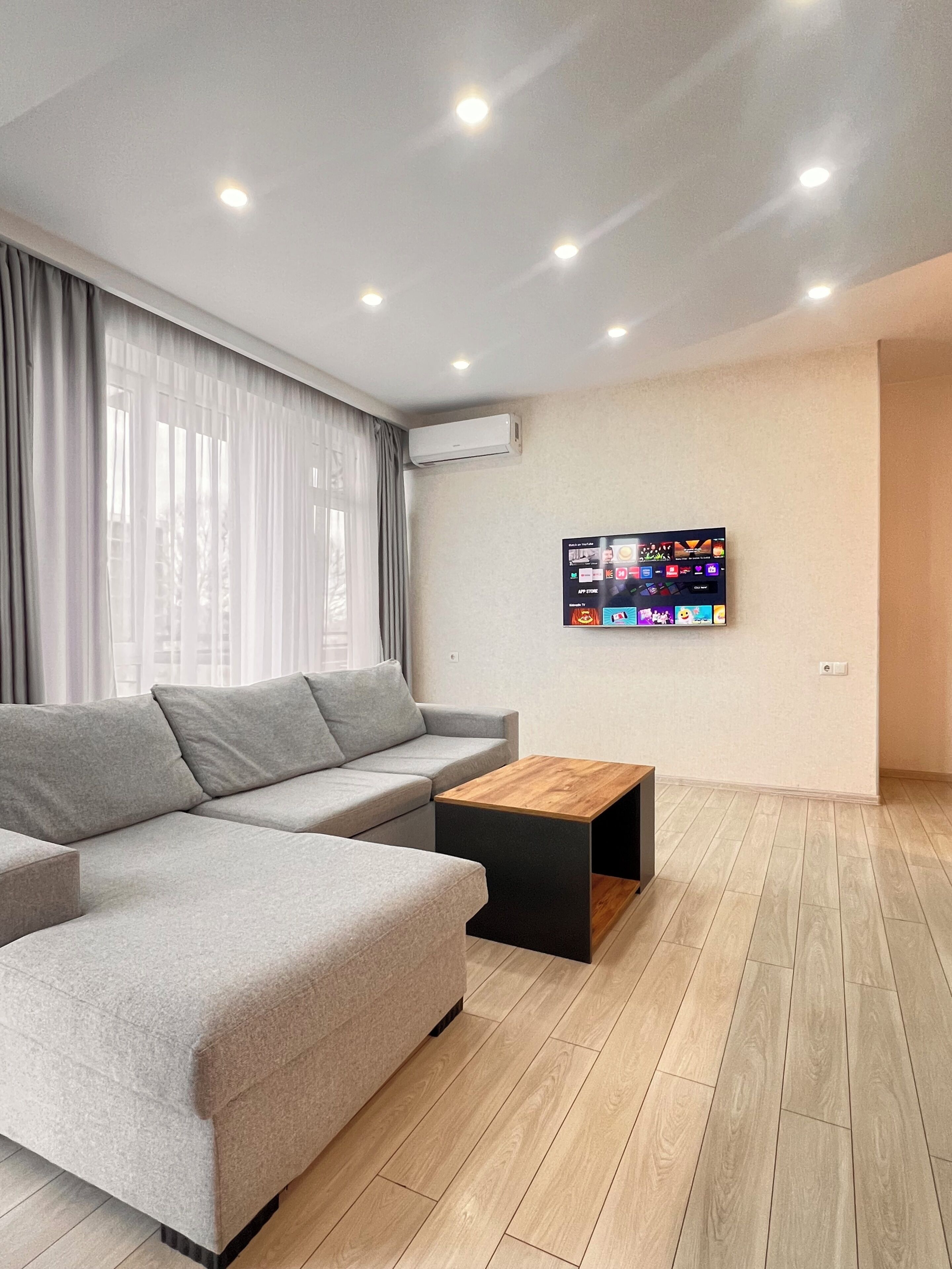 Uma sala de estar moderna com um sofá seccional cinza, uma mesa de centro de madeira e uma TV de tela plana na parede.