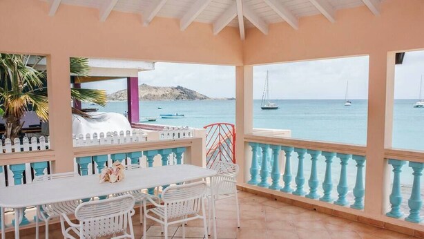 Un espace extérieur abrité, avec une table et des chaises, donnant sur une mer calme où naviguent des bateaux.