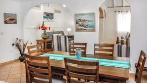 Un coin repas avec une table en bois, des chaises et un vase de fleurs. Les murs sont ornés de tableaux et, à travers une arche, on aperçoit la mer.