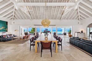 A spacious living area with a large wooden dining table surrounded by chairs, a black leather sofa, and a view of the ocean through large windows. - Petite Plage 5   Exclusive Beachfront Villa (Philipsburg)