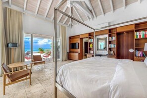 A hotel room with a large bed, a desk, a chair, a TV, and a bookshelf. There is a balcony with a view of the ocean. - Petite Plage 5   Exclusive Beachfront Villa (Philipsburg)