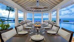 Un área de comedor cubierta con vistas al océano, columnas blancas y una mesa central dispuesta para una comida.