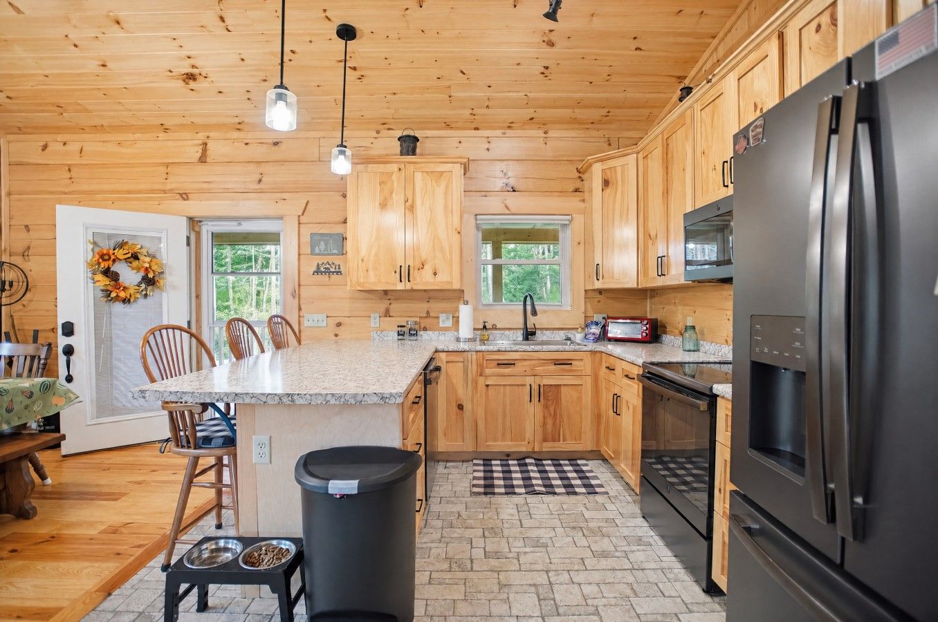 A kitchen with wooden cabinets, a granite countertop, a stainless steel refrigerator, and a black trash can.