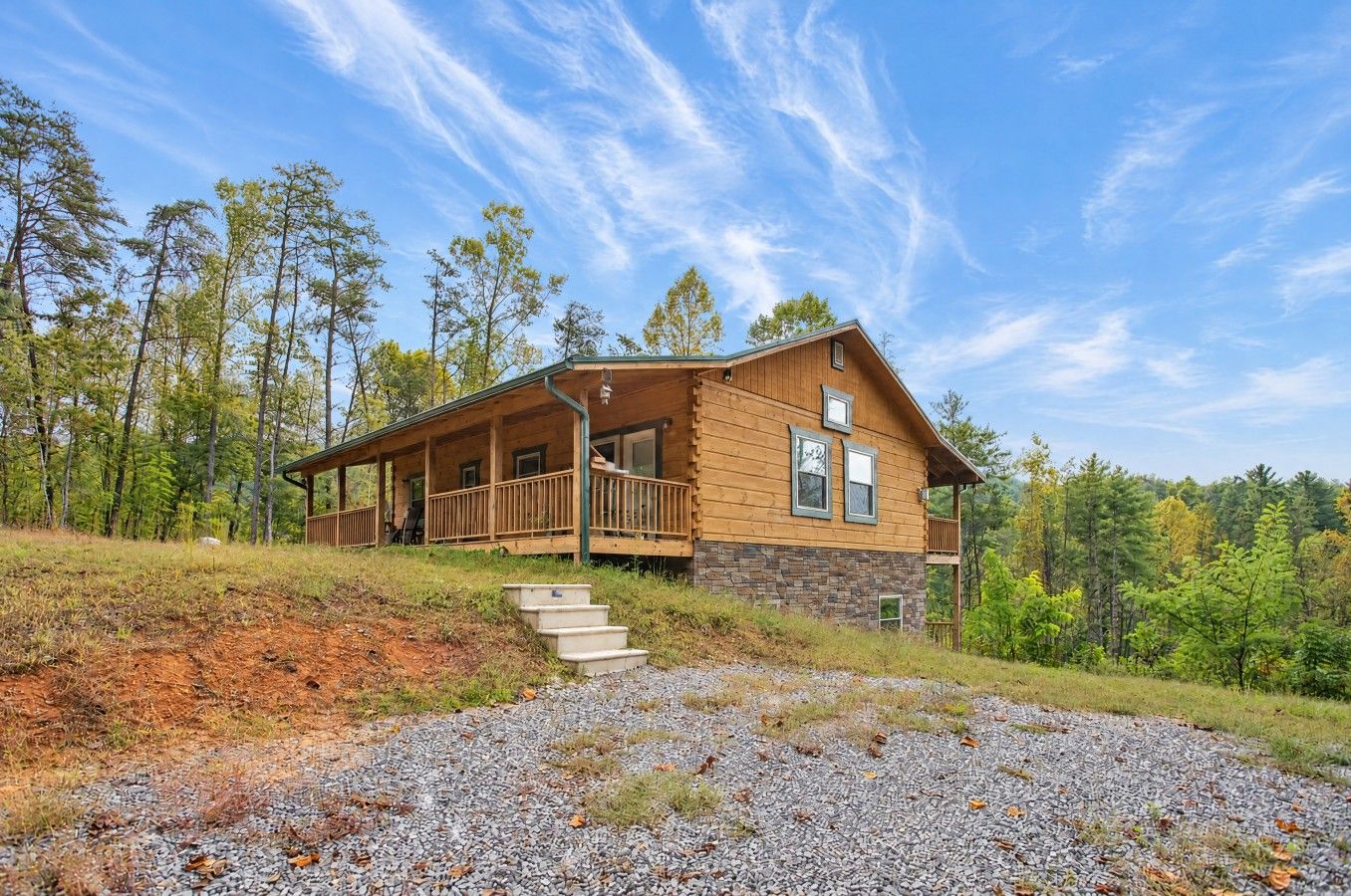 A wooden cabin with a porch, stone foundation, and a gravel driveway.