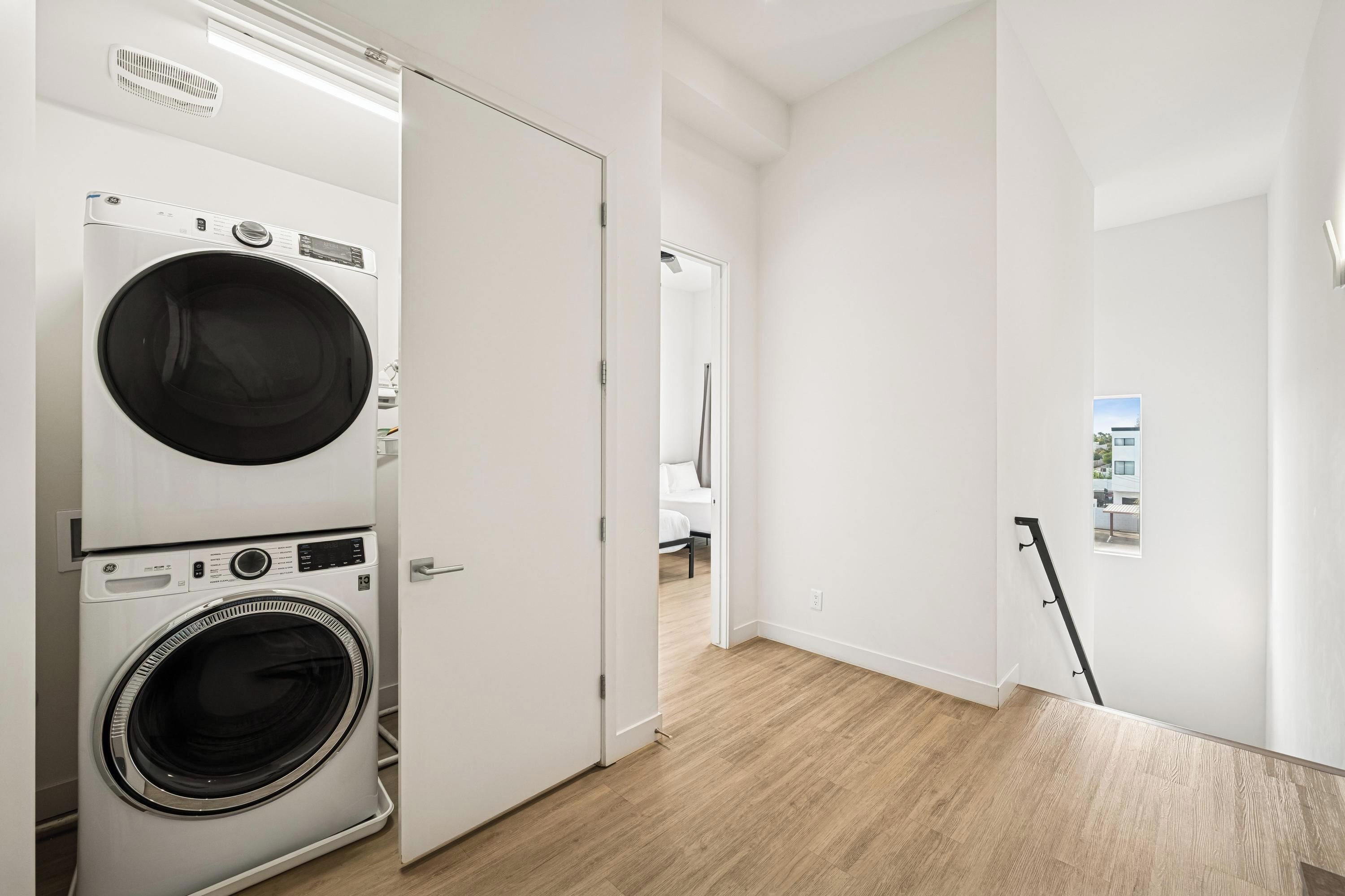 Un lavadero con lavadoras y secadoras apiladas, suelo de madera y vista a un baño con ducha.