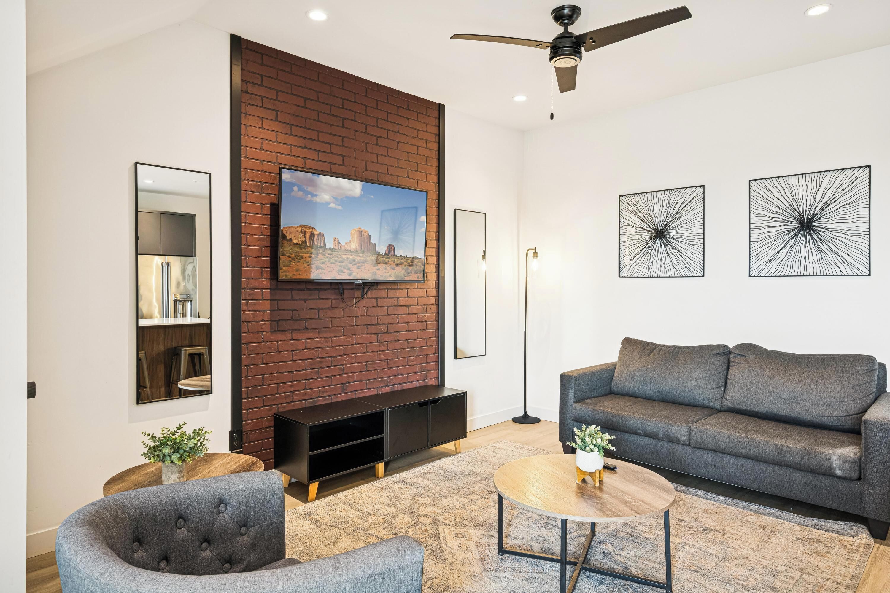 Una sala moderna con una pared de ladrillo decorativa, un televisor de pantalla plana, un sofá gris, una mesa de centro y un ventilador de techo.