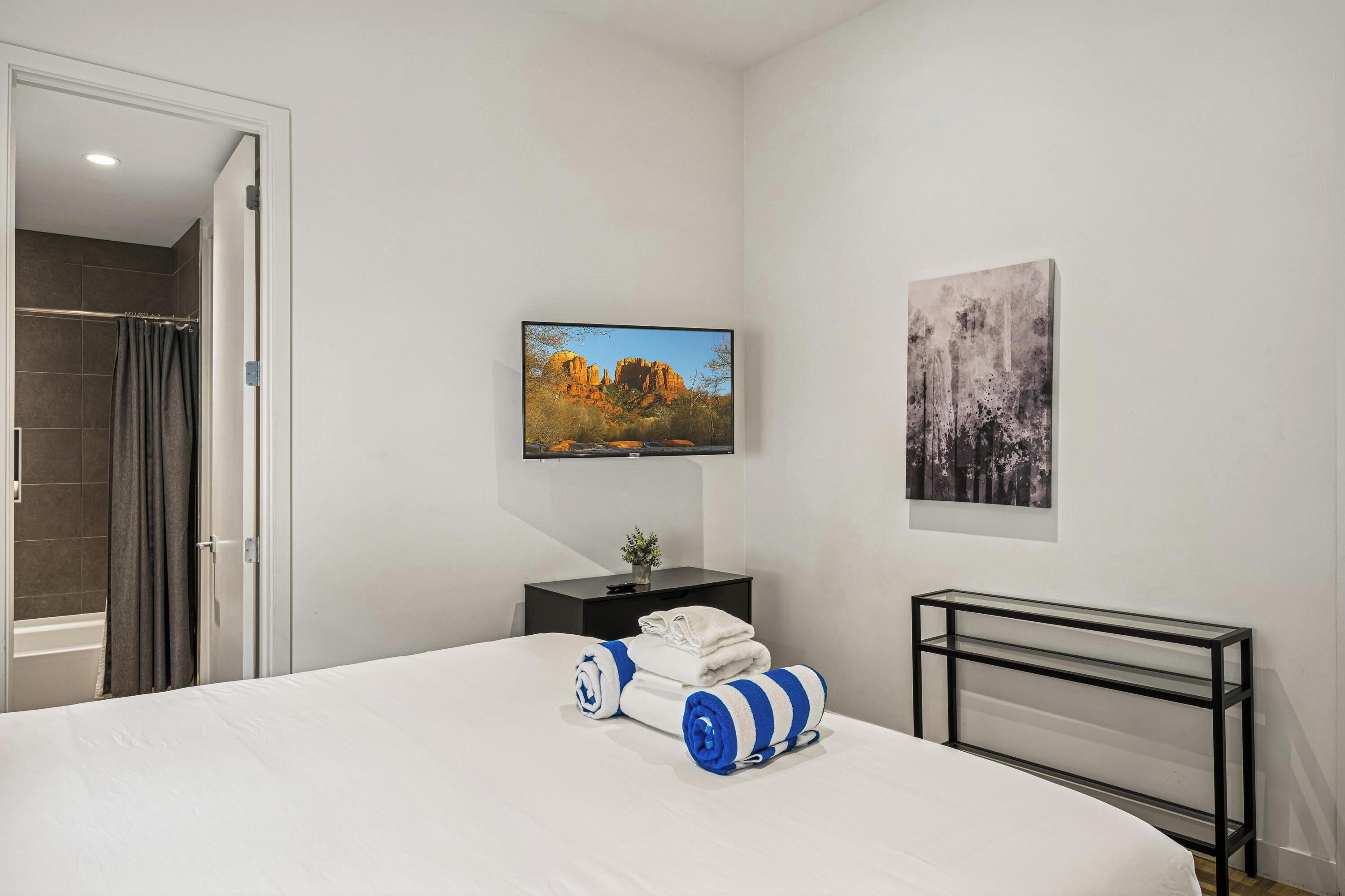 A hotel room with a bed, a TV mounted on the wall, a painting, and a small table with a plant.