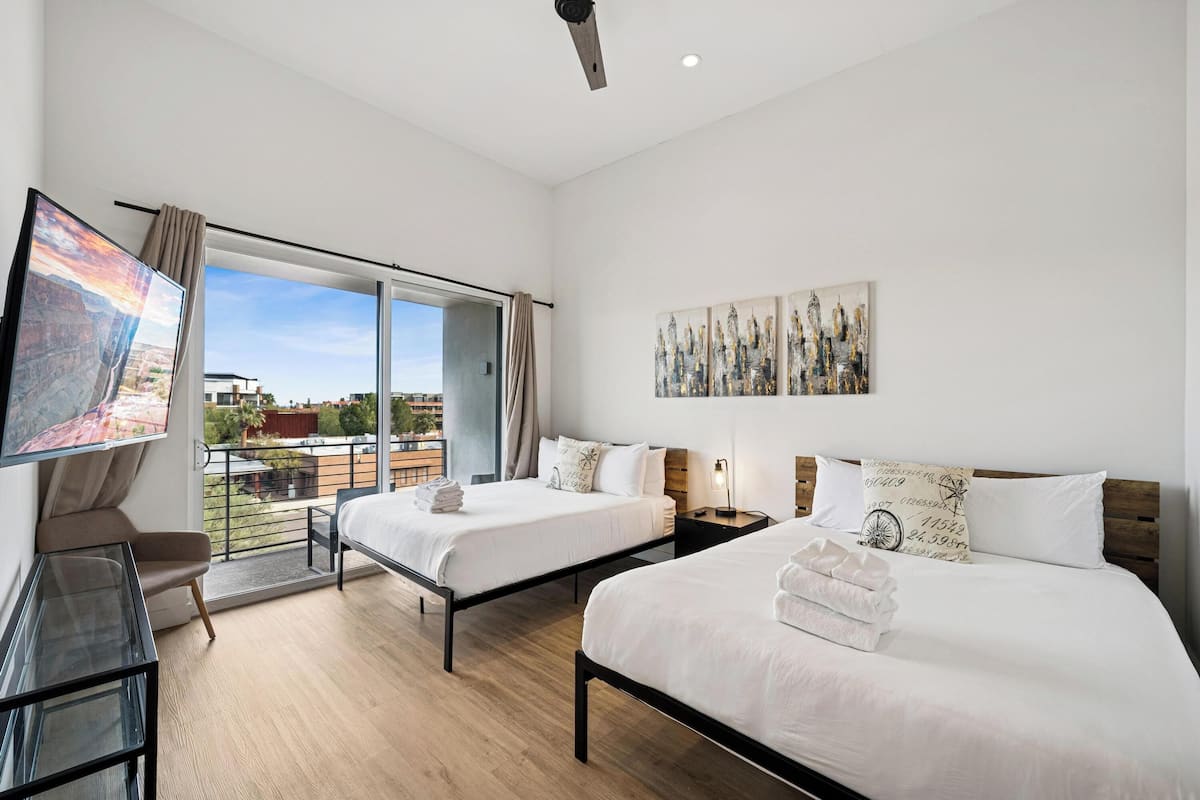 A hotel room with two beds, a TV, a desk, and a balcony with a city view.