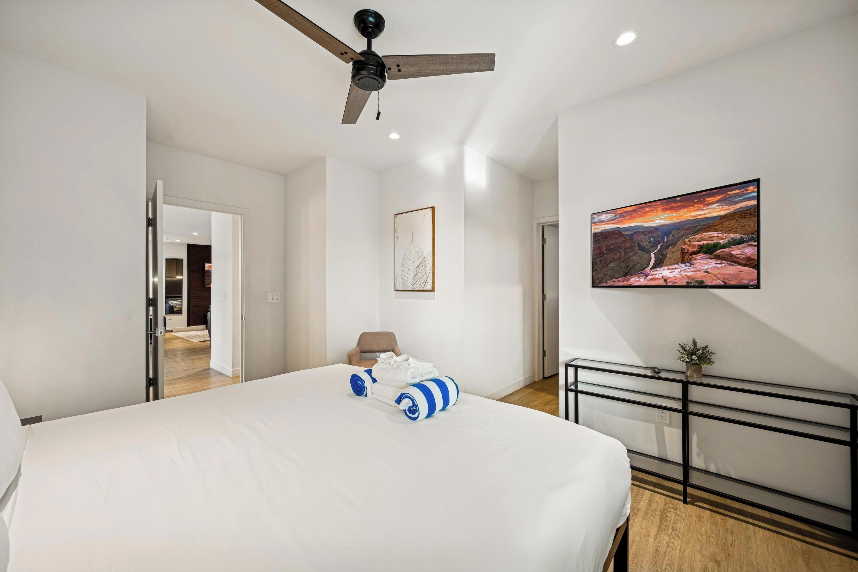 Une chambre moderne avec un grand lit, un ventilateur de plafond, un tableau représentant un canyon, et une vue sur la cuisine et la salle de bain à travers une porte ouverte.