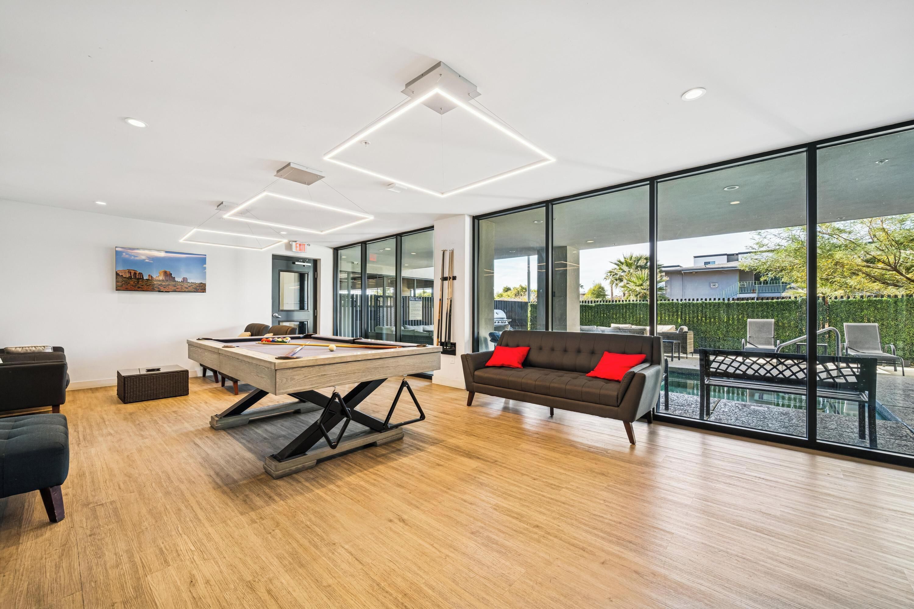 A modern lounge area with a pool table, a sofa with red cushions, and large windows offering a view of the outdoors.
