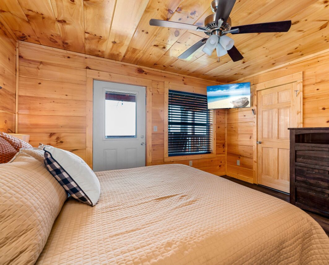 A wooden-paneled bedroom with a bed, a ceiling fan, a door, and a window with blinds.