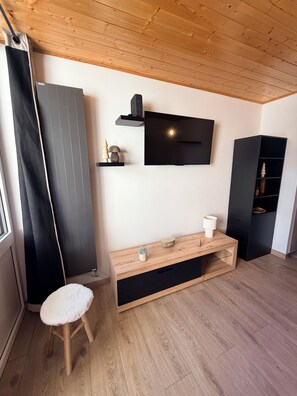 Ein modernes Zimmer mit Holzboden, einem an der Wand montierten schwarzen Fernseher, einem Holztischelement, einem weißen gepolsterten Hocker und einem schwarzen Schrank.