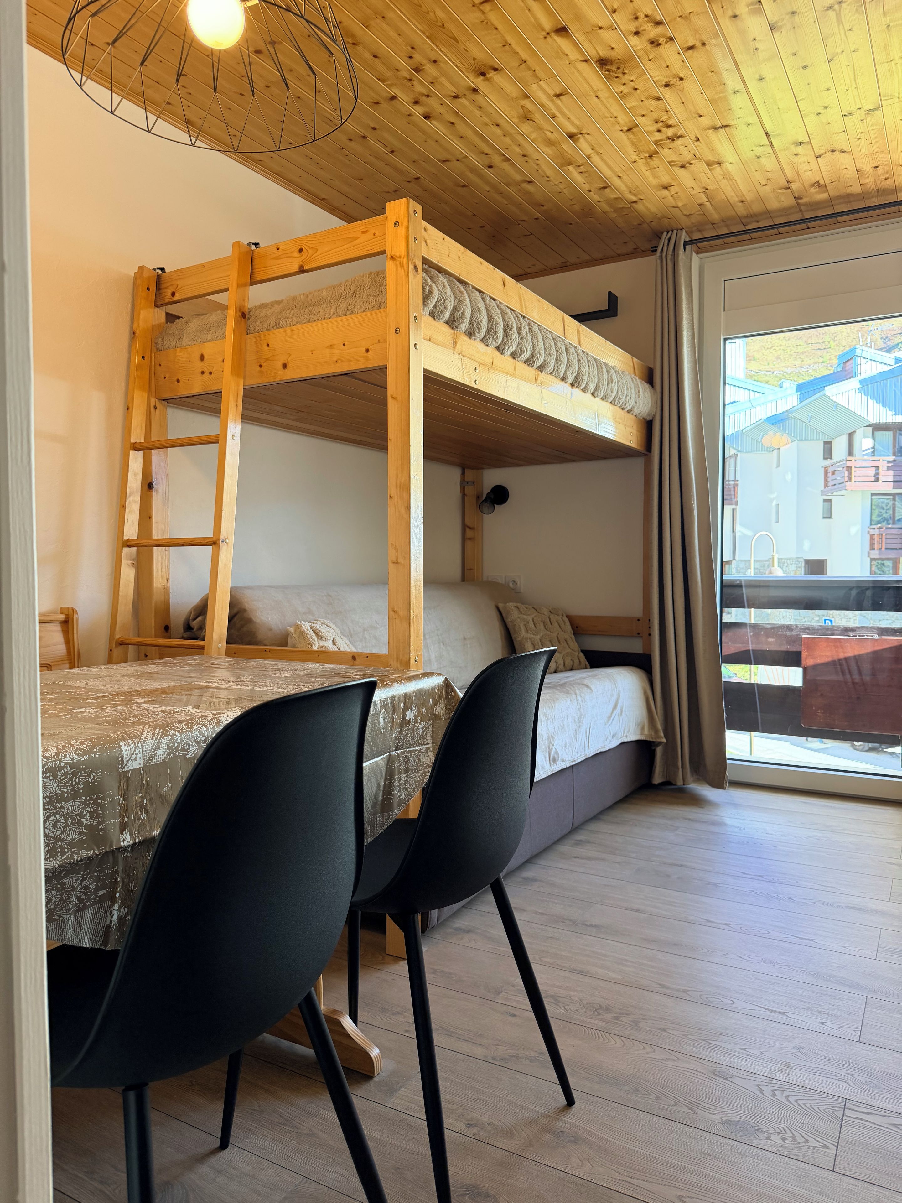 Un lit superposé avec une échelle, une table avec des chaises et une fenêtre donnant sur des immeubles.