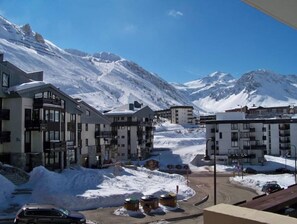 Un pueblo nevado al pie de una montaña, con edificios modernos, cielo despejado y una calle con automóviles estacionados.