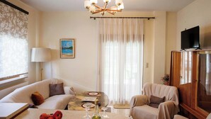 A living room with a sofa, armchairs, a coffee table with a bottle and glasses, a TV on a cabinet, and a painting on the wall. - Anastasia s Kastoria Cozy Modern Garden Retreat (Kastoria)