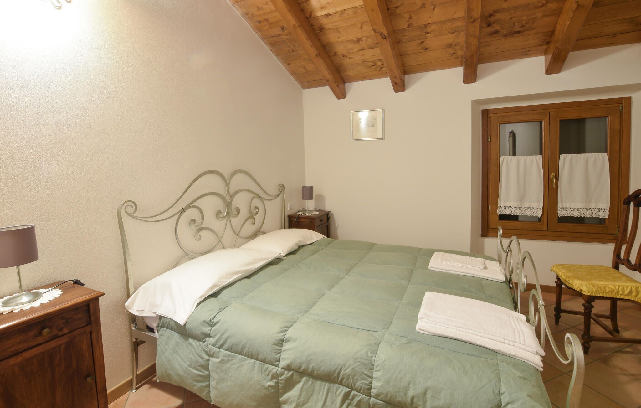 Een slaapkamer met een houten plafond, een bed met een groene sprei, een nachtkastje met een lamp, een houten stoel en een raam met gordijnen.