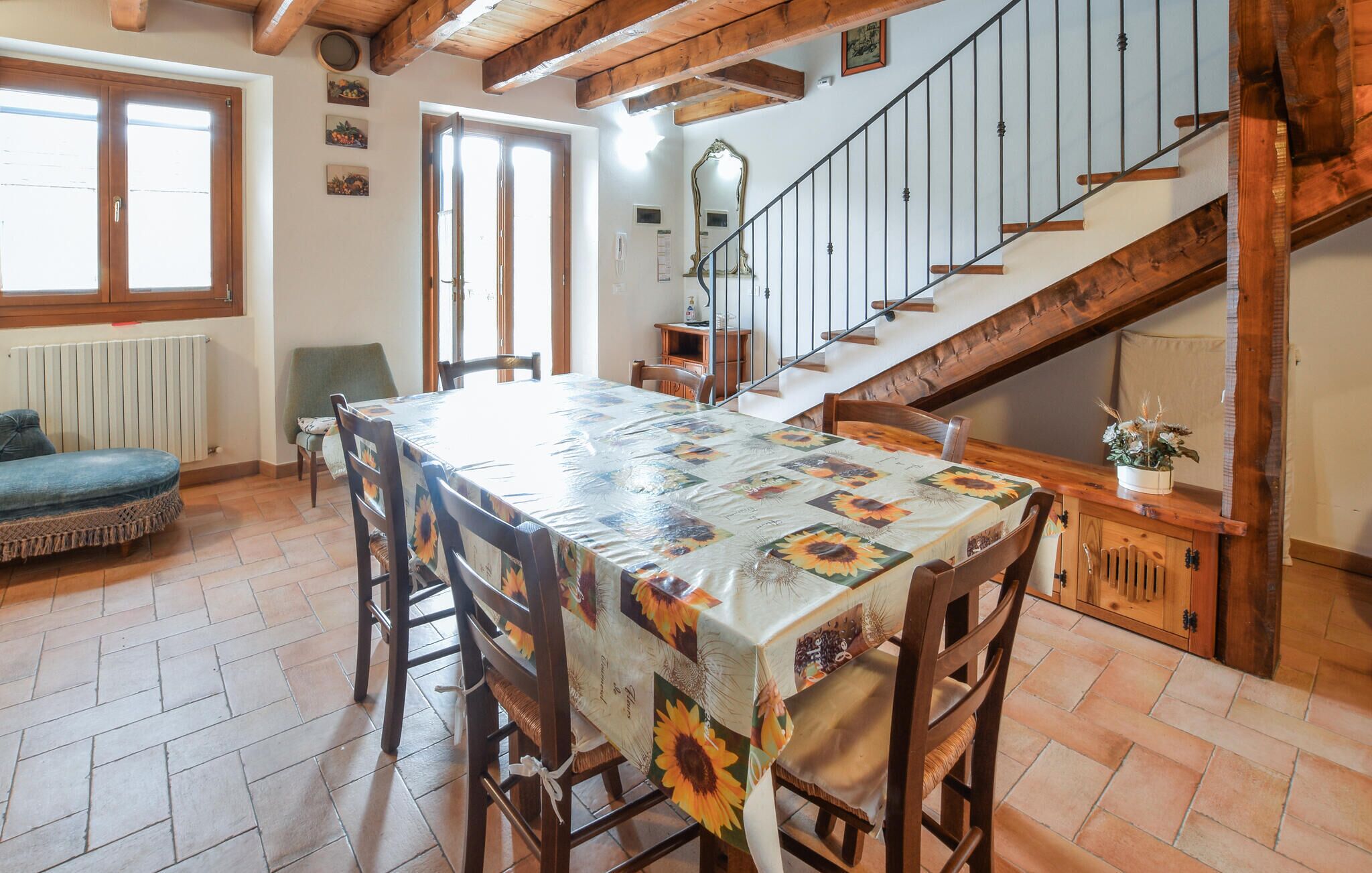 Ein Essbereich mit einem Tisch, bedeckt mit einer Sonnenblumen-Tischdecke, Holzstühlen, einer Couch und einer Treppe, die zu einer oberen Ebene führt.