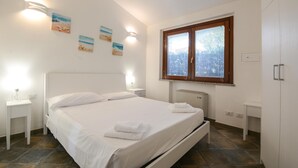 A neatly arranged bedroom with a bed, bedside tables, a window with blinds, and wall-mounted artwork. - Cozy home in Costa Rei (Costa Rei)