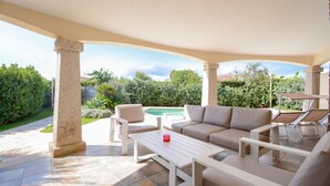 An outdoor patio with a beige sofa, white coffee table, and lounge chairs. A swimming pool is visible in the background. - Cozy home in Costa Rei (Costa Rei)