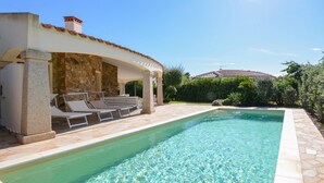 A swimming pool with clear water, a covered seating area with lounge chairs, and a stone wall. - Cozy home in Costa Rei (Costa Rei)