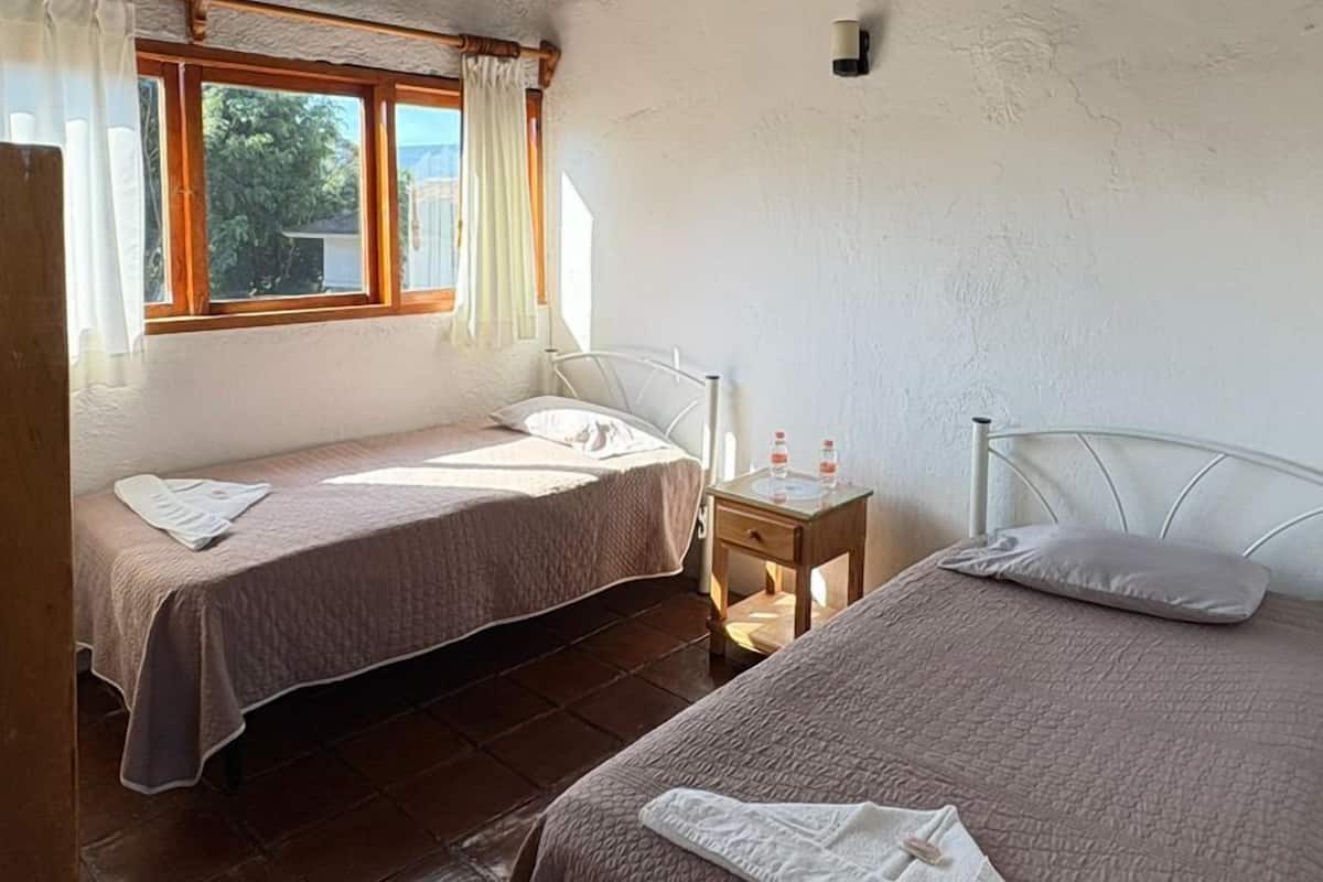 A room with two beds, a window with curtains, a wooden ceiling, and a small table with a water bottle.