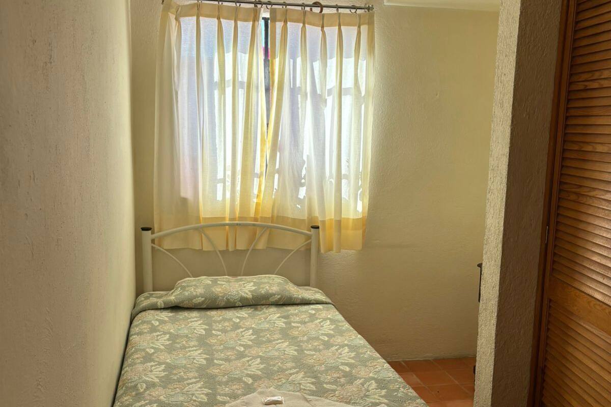 A small, single-bed room with a floral-patterned bedspread, a window with sheer curtains, and a wooden door.