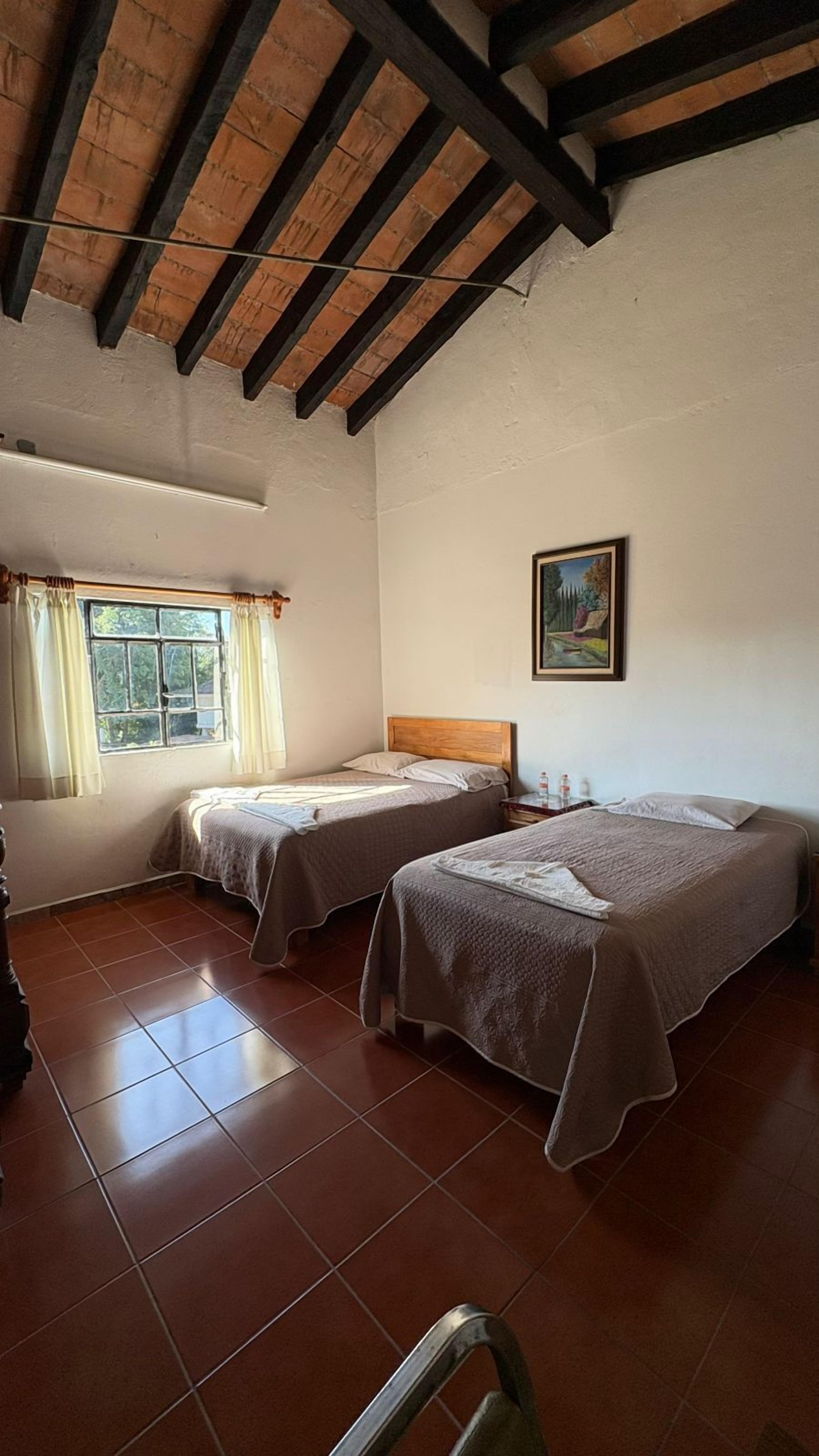A room with two single beds, a window with curtains, a painting on the wall, and a tiled floor.