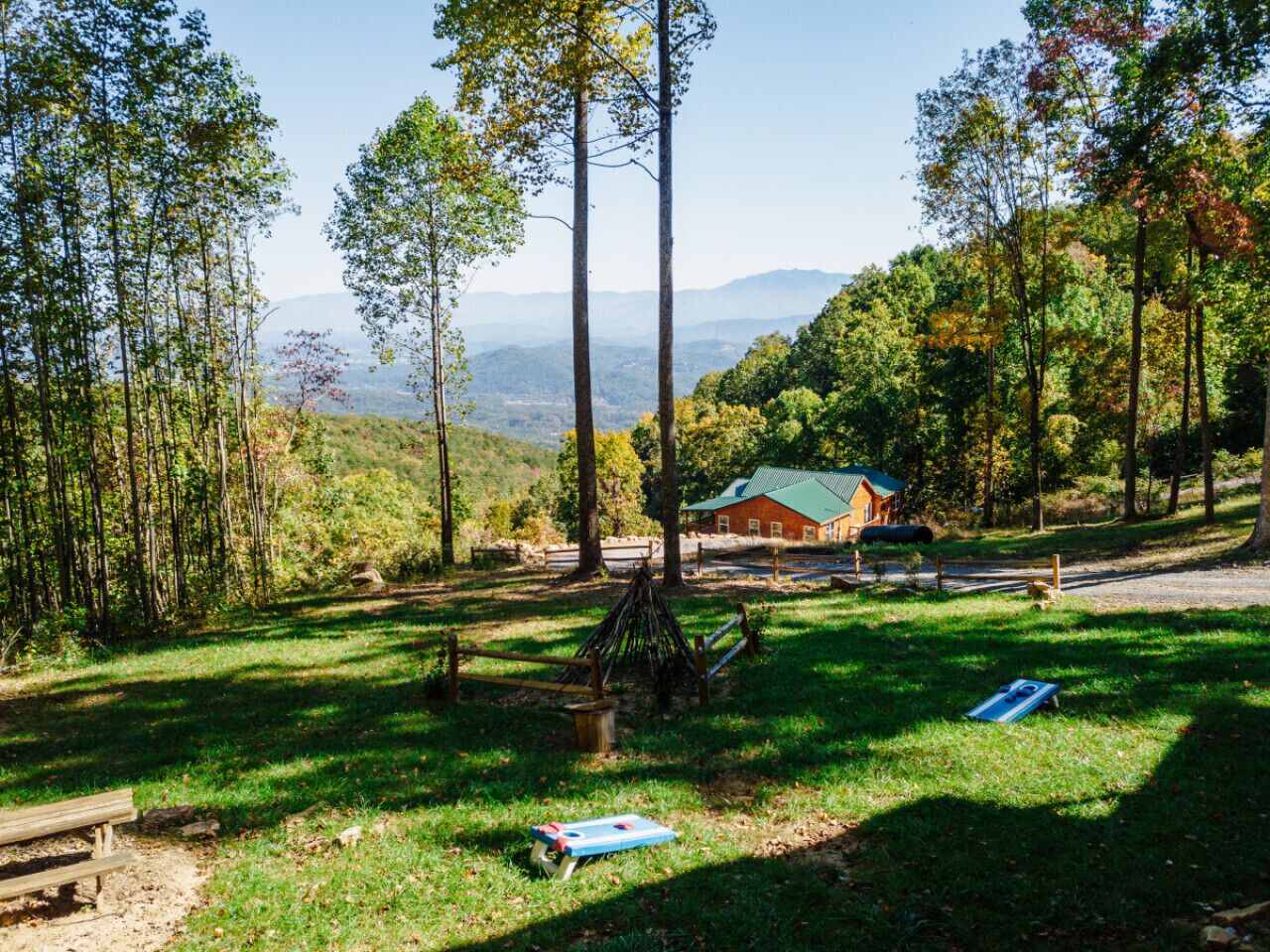 A rustic cabin with a green roof surrounded by a forested area with tall trees and a clear view of distant mountains.