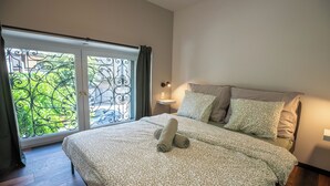Een slaapkamer met een bed, nachtkastje, lamp en uitzicht door een raam met balkon.