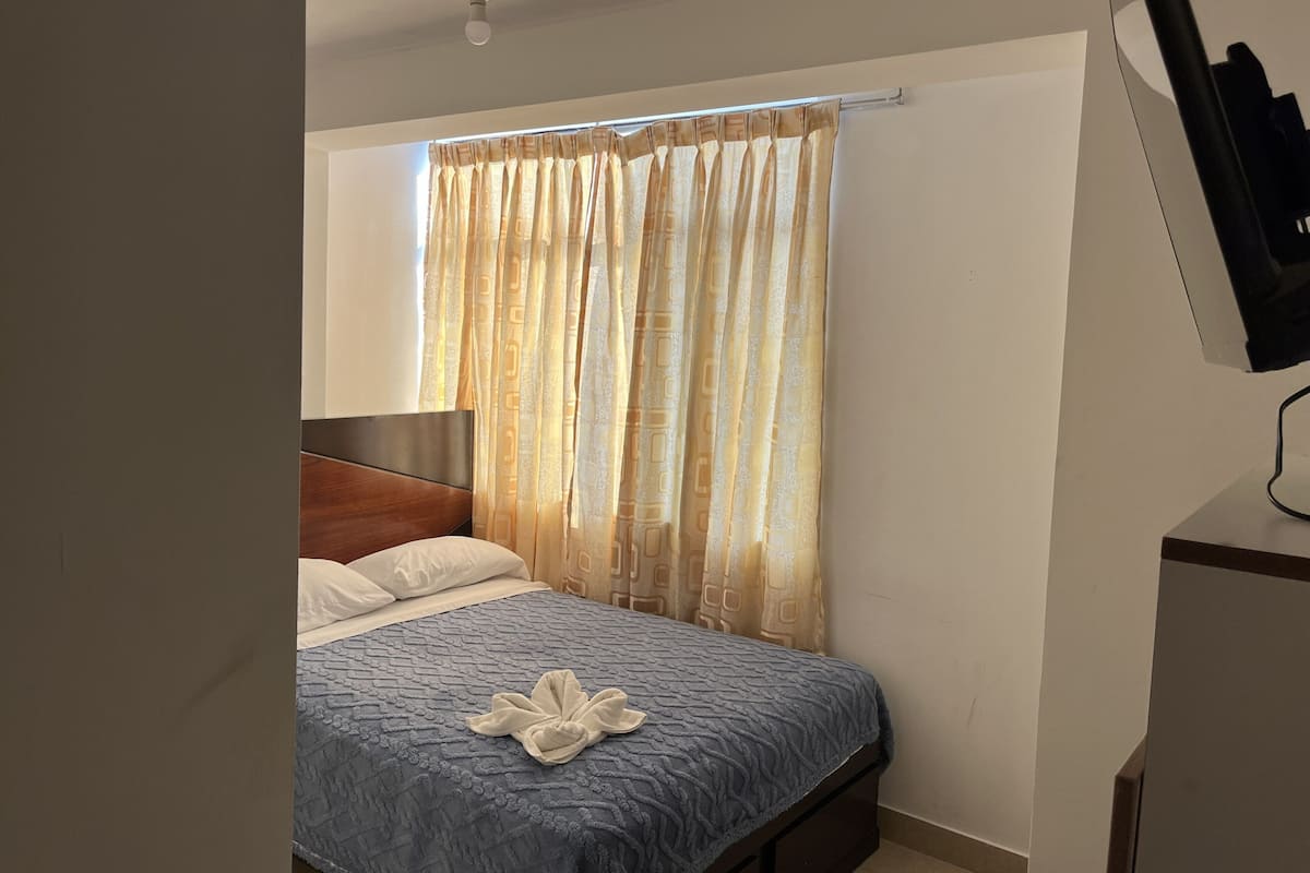 A bedroom with a bed, a headboard, a window with curtains, and a television on a stand.