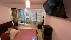 A hotel room with a bed, a TV, a desk, and a bathroom visible through a glass door. - hostal milagros (Chancay)