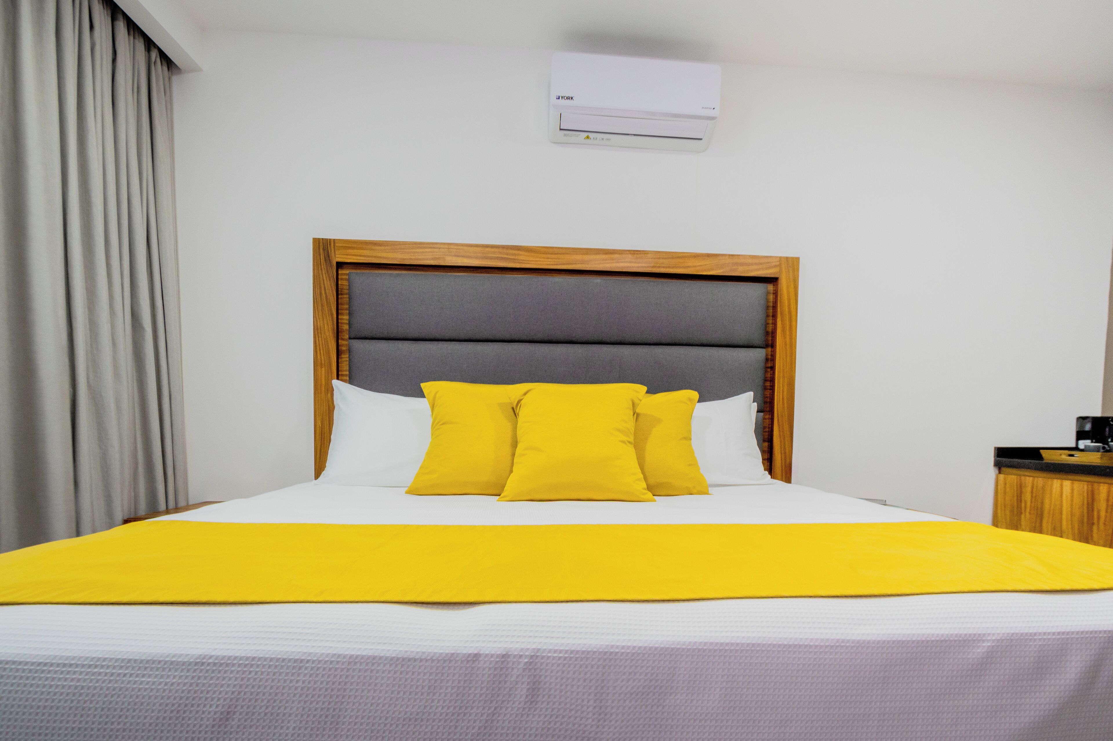 Una camera d'hotel con un letto, una testiera in legno, una parete bianca, un condizionatore d'aria e un comodino con una macchina per il caffè.
