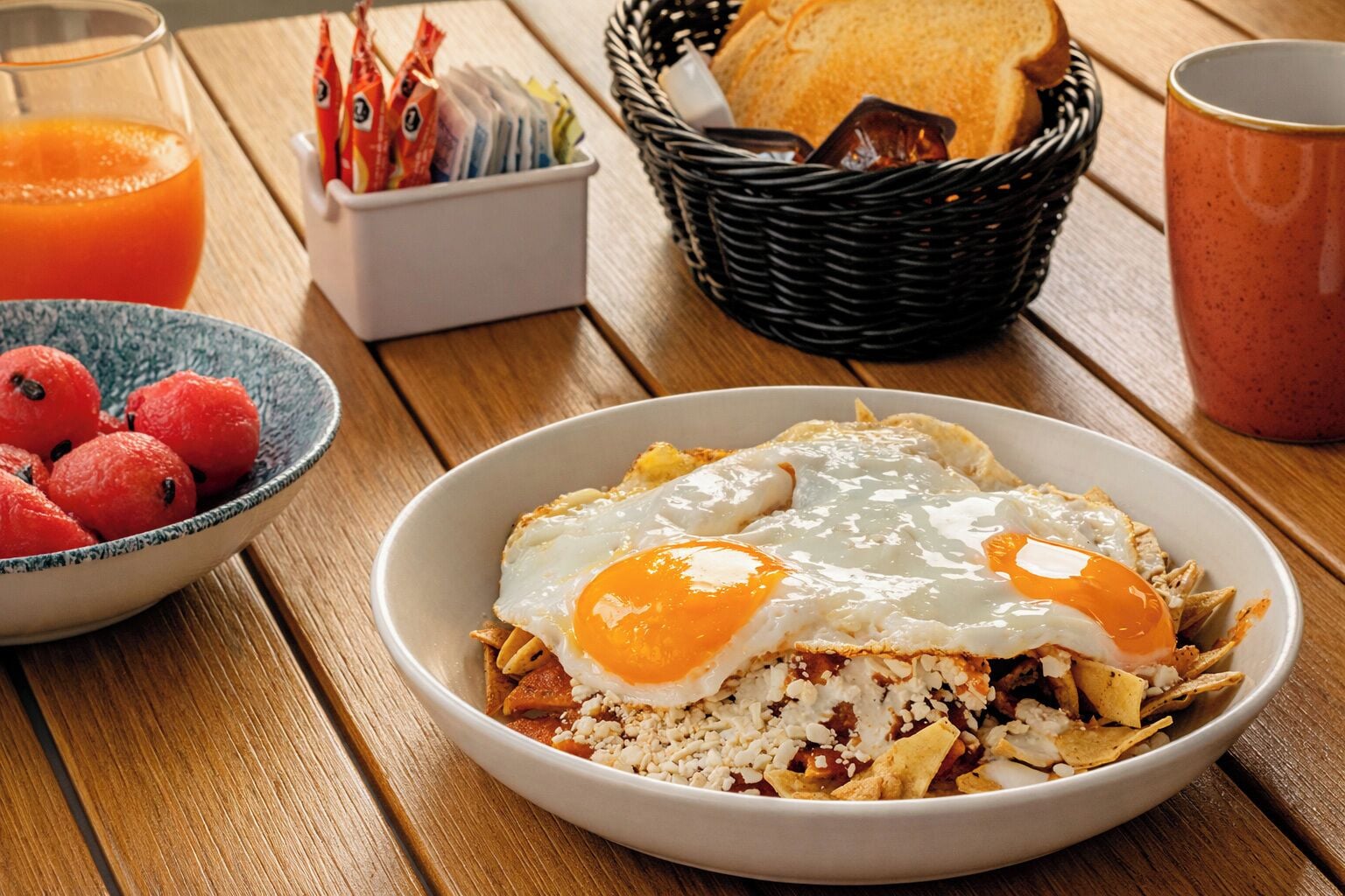 A breakfast plate with eggs, a bowl of fruit, and a basket of bread on a wooden table.