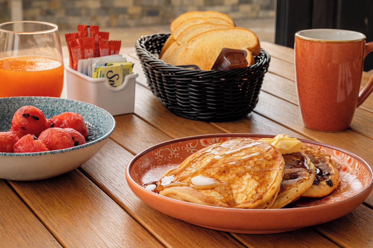 A breakfast setting with pancakes, a bowl of berries, a basket of bread, a mug, and a glass of orange juice.