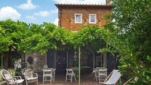 A patio with white metal chairs and tables, surrounded by lush green trees and a brick building in the background. - Amazing home in Firenze (Firenze)