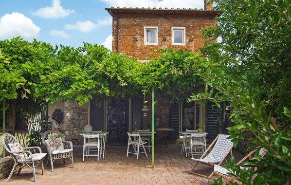 A patio with white metal chairs and tables, surrounded by lush green trees and a brick building in the background. - Amazing home in Firenze (Firenze)