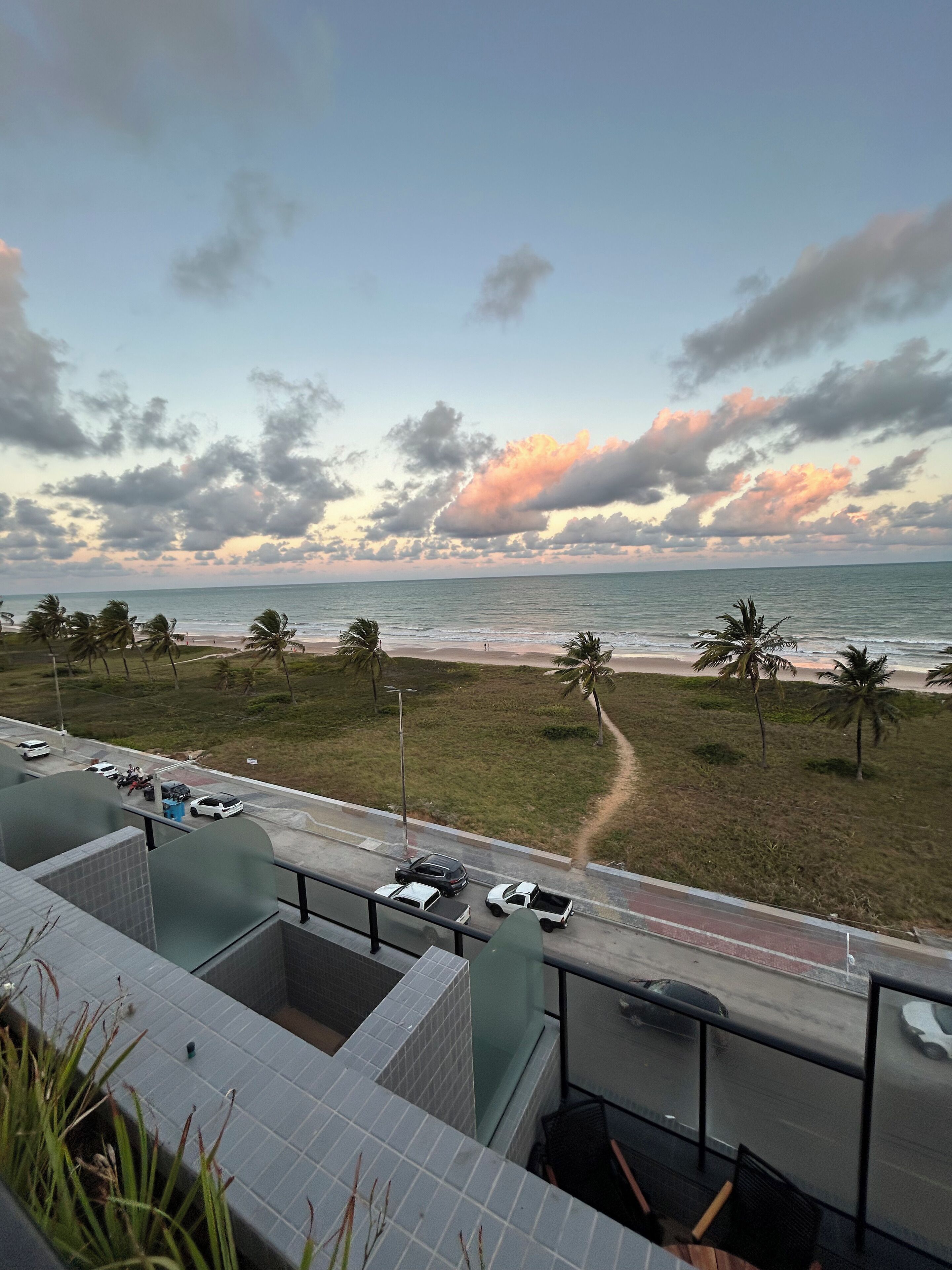 A view of a beach from a building with a tiled roof, a road with parked cars, and palm trees.